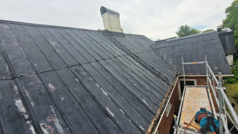 Standing-seam metal roof with visible wear and scaffolding at the eaves in Brantevik.