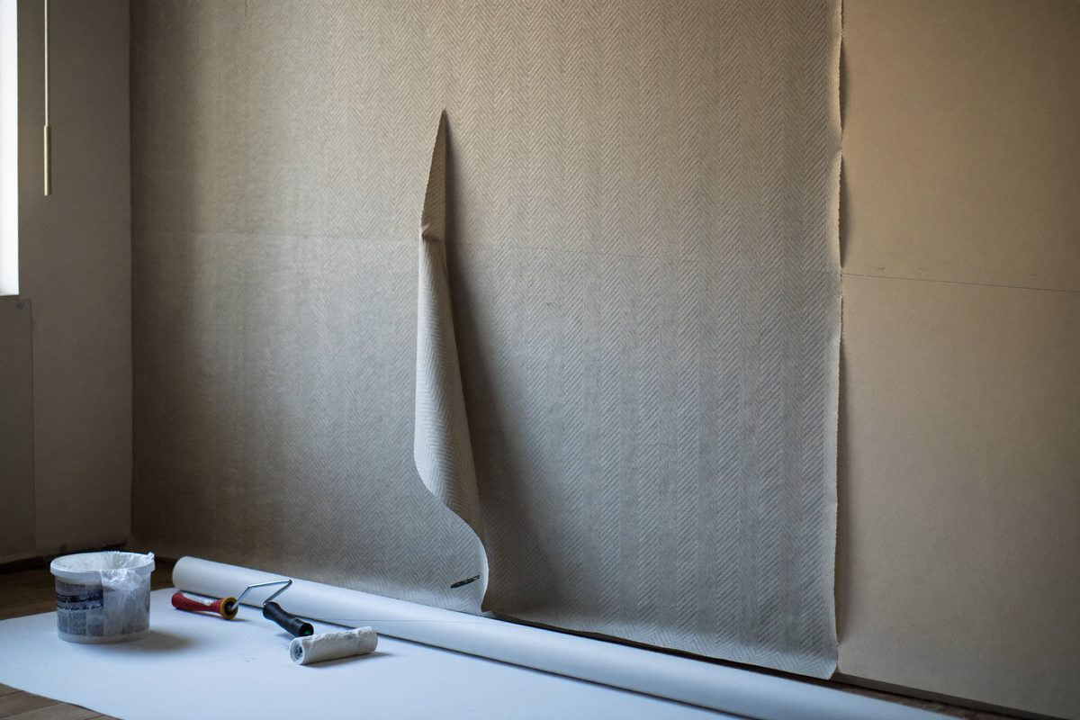 Vägg under tapetsering där två våder sitter uppe och nästa våd ligger delvis utrullad, med klisterhink, roller och sömroller på golvet