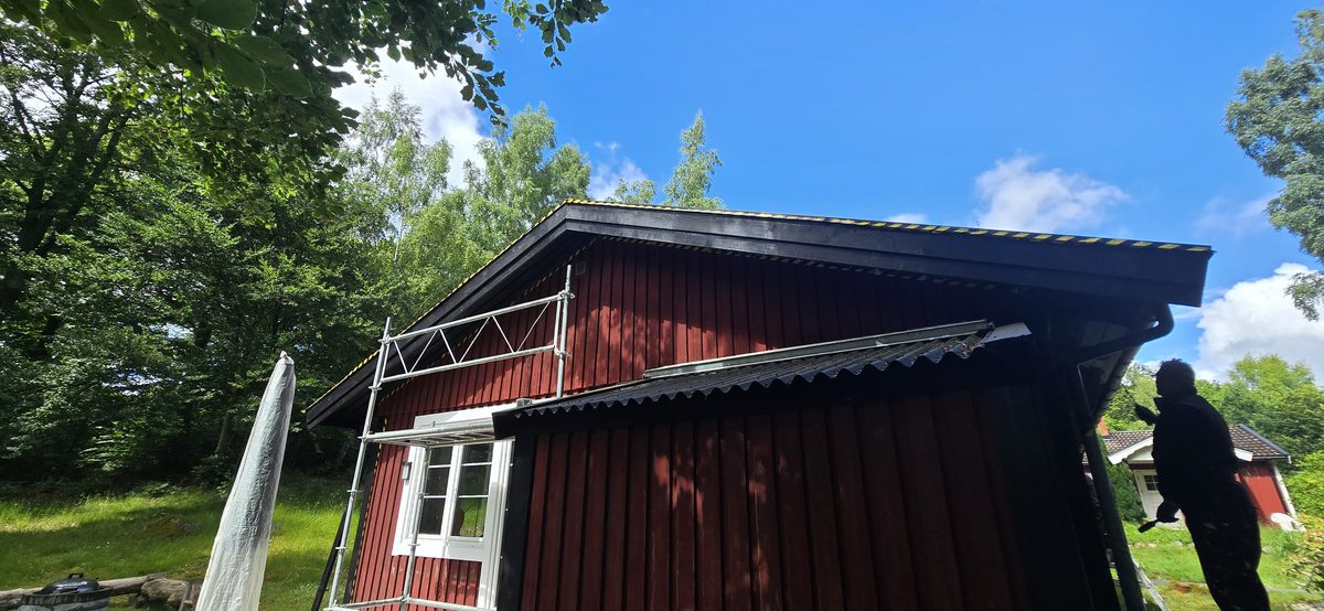 Målare i arbete på ställningen vid det rödmålade trähuset i Lycke, med svart korrugerat tak ovanför
