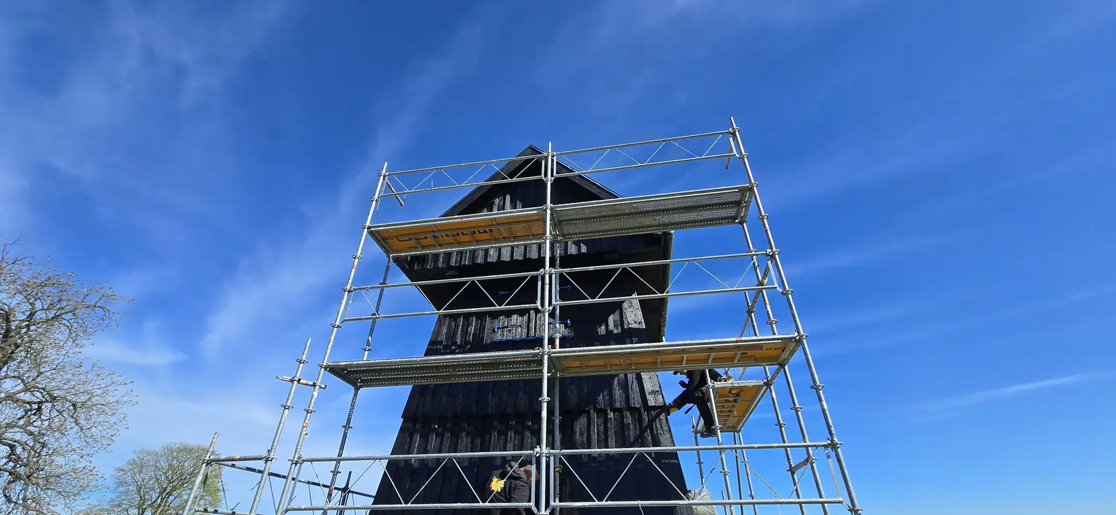 Mörk klockstapel vid Kvidinge kyrka omgiven av ställning
